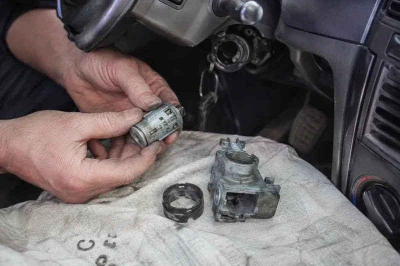 Hands holding a removed ignition cylinder beside a car steering column, with the housing and small parts on a cloth. — Ignition Lock Repair, Woodstock
