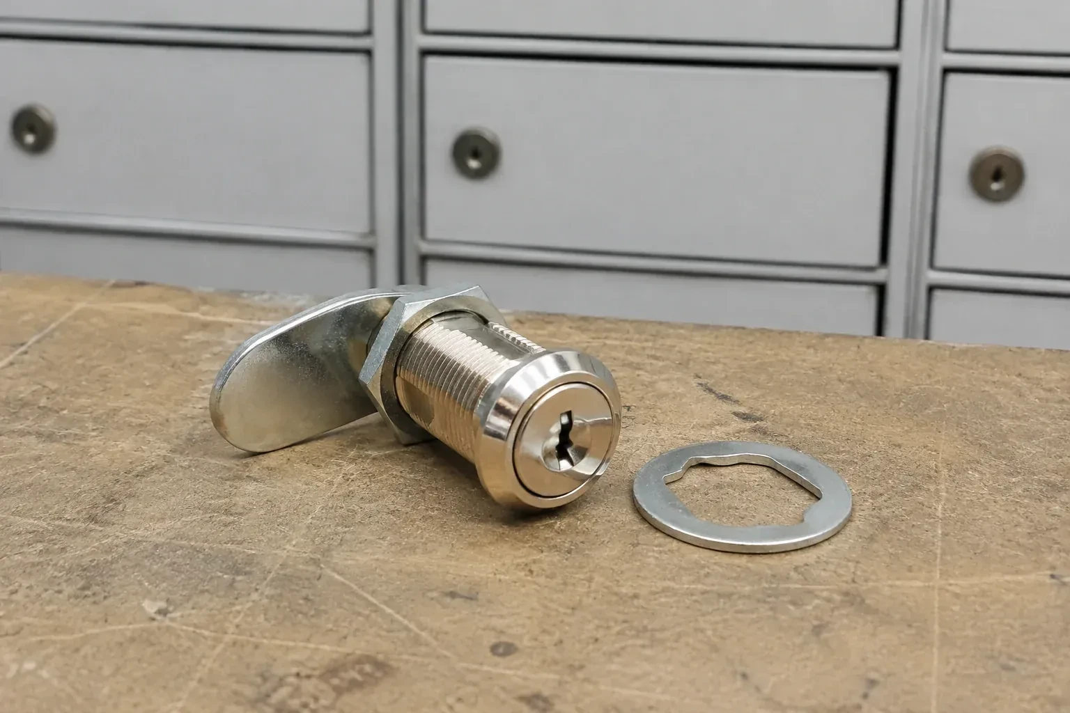 Detached threaded cam lock and mounting washer on a workbench in front of closed mail compartments. — Mailbox Lock Replacement, Woodstock